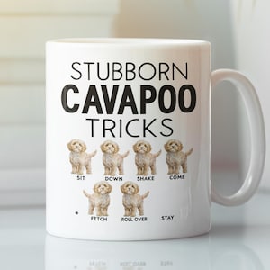 Op de afbeelding: Witte keramische mok met een zwarte afbeelding waarop "Stubborn Cavapoo Tricks" staat. De afbeelding bevat zes illustraties van cavapoos die trucs uitvoeren, waaronder zitten, liggen, schudden, komen, apporteren, rollen en blijven.