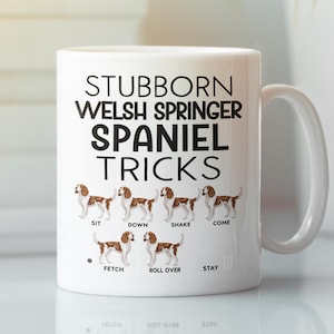 May include: White ceramic mug featuring the text "STUBBORN WELSH SPRINGER SPANIEL TRICKS" with illustrations of a dog performing tricks. The tricks include sit, down, shake, come, fetch, roll over, and stay.