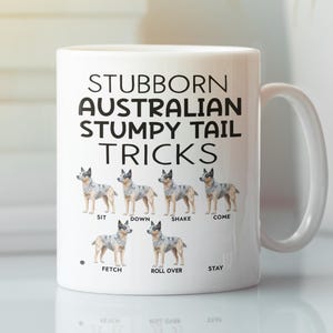 Può includere: Tazza in ceramica bianca con la scritta "STUBBORN AUSTRALIAN STUMPY TAIL TRICKS" e illustrazioni di cani Australian Stumpy Tail che eseguono trucchi. La tazza mostra cani che fanno trucchi come seduto, giù, scuoti e vieni. Ha un manico curvo.