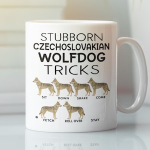 May include: White ceramic mug featuring the text "STUBBORN CZECHOSLOVAKIAN WOLFDOG TRICKS" in black. Below the text are illustrations of wolfdogs demonstrating tricks like sit, down, shake, come, fetch, roll over, and stay. The mug has a white handle.