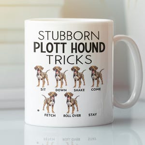 May include: White ceramic coffee mug featuring a black and white illustration of a Plott Hound dog. The mug displays the text "Stubborn Plott Hound Tricks" and lists various dog tricks with an illustration of the dog performing each trick.