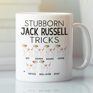 May include: White ceramic mug featuring the text "STUBBORN JACK RUSSELL TRICKS" with illustrations of a Jack Russell Terrier demonstrating tricks. The tricks include "SIT", "DOWN", "SHAKE", "COME", "FETCH", "ROLL OVER", and "STAY".