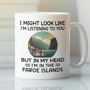 May include: White ceramic mug with a handle featuring the text "I MIGHT LOOK LIKE I'M LISTENING TO YOU BUT IN MY HEAD I'M IN THE FAROE ISLANDS." The mug has a circular image of a coastal village with mountains and water.