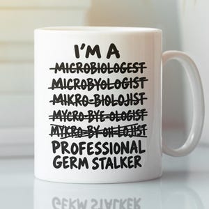 Könnte beinhalten: Ein weißer Keramikbecher mit schwarzem Text, der lautet: I'M A, gefolgt von mehreren falsch geschriebenen Versionen des Wortes microbiologist, die jeweils mit einer horizontalen Linie durchgestrichen sind, und endet mit PROFESSIONAL GERM STALKER.