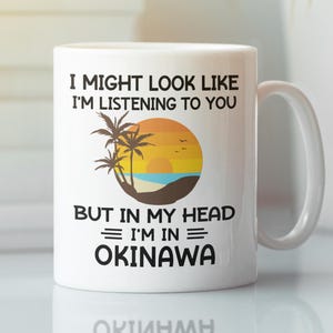 May include: White ceramic mug with a handle. The mug features the text "I MIGHT LOOK LIKE I'M LISTENING TO YOU BUT IN MY HEAD I'M IN OKINAWA" with a sunset and palm tree graphic. The mug is a standard 11 oz size.