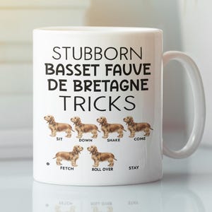 Puede incluir: Taza de cerámica blanca con el texto "STUBBORN BASSET FAUVE DE BRETAGNE TRICKS" en negro. La taza presenta ilustraciones de un perro Basset Fauve de Bretagne realizando trucos. La taza tiene un asa blanca y es adecuada para bebidas calientes.