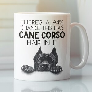 Könnte beinhalten: Weiße Keramiktasse mit einer schwarzen Cane Corso Hunde-Grafik und dem Text "THERE'S A 94% CHANCE THIS HAS CANE CORSO HAIR IN IT". Die Tasse hat einen gebogenen Henkel. Der Hintergrund ist unscharf.
