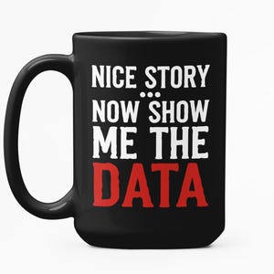 May include: A black ceramic mug with a large handle. The mug displays the white text "NICE STORY NOW SHOW ME THE DATA", with the word "DATA" in red. The mug is set against a white background.