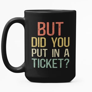 Könnte beinhalten: Schwarze Kaffeetasse mit dem Text "BUT DID YOU PUT IN A TICKET?" in einer Retro-Schriftart. Der Text ist in Orange, Gelb und Türkis gehalten. Die Tasse hat einen großen Henkel.