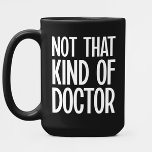 Könnte beinhalten: Eine schwarze Keramik-Tasse mit großem Henkel. Die Tasse zeigt den weißen Text "NOT THAT KIND OF DOCTOR" in einer fetten, serifenlosen Schrift. Die Tasse ist für Getränke konzipiert und eignet sich für den Heim- oder Bürogebrauch.