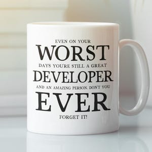 Puede incluir: Taza de cerámica blanca con texto negro: "EVEN ON YOUR WORST DAYS YOU'RE STILL A GREAT DEVELOPER AND AN AMAZING PERSON. DON'T YOU EVER FORGET IT!". La taza tiene un asa curva.