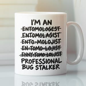 Op de afbeelding: Een witte keramische mok met een handvat en zwarte tekst: I'M AN ENTOMOLOGIST, met verschillende doorgestreepte spelfouten van het woord, gevolgd door de tekst PROFESSIONAL BUG STALKER in dikgedrukte hoofdletters.