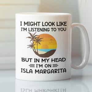May include: White ceramic mug with a handle featuring the text "I MIGHT LOOK LIKE I'M LISTENING TO YOU BUT IN MY HEAD I'M ON ISLA MARGARITA." The mug has a beach scene with palm trees, a sunset, and the ocean.