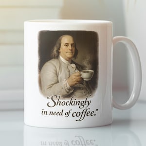 Könnte beinhalten: Weiße Keramik-Kaffeetasse mit einem Porträt von Benjamin Franklin, der eine Kaffeetasse hält. Auf der Tasse steht der Spruch "Shockingly in need of coffee." in schwarzer Schrift. Die Tasse hat einen weißen Henkel. Fassungsvermögen ca. 325 ml.