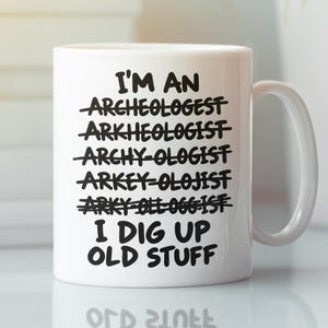 Puede incluir: Una taza de cerámica blanca con texto negro que dice: I'M AN ARCHEOLOGIST, seguido de varias variaciones mal escritas tachadas, y termina con la frase I DIG UP OLD STUFF en letras mayúsculas negras, claras y en negrita.