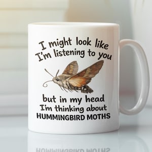 Op de afbeelding: Witte keramische mok met een illustratie van een kolibrievlinder en een humoristische quote. De mok zegt: "I might look like I'm listening to you but in my head I'm thinking about HUMMINGBIRD MOTHS."