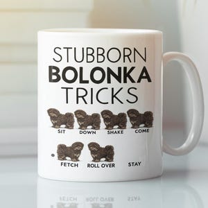 May include: White ceramic mug with the text "STUBBORN BOLONKA TRICKS" and illustrations of a Bolonka dog performing tricks. The tricks include sit, down, shake, come, fetch, roll over, and stay.