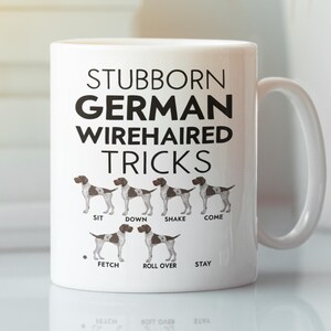 May include: White ceramic mug featuring the text "STUBBORN GERMAN WIREHAIRED TRICKS" in black. Below the text are illustrations of German Wirehaired Pointers demonstrating commands like "SIT", "DOWN", "SHAKE", "COME", "FETCH", "ROLL OVER", and "STAY".