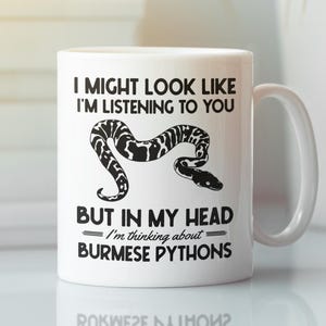 Könnte beinhalten: Weiße Keramik-Tasse mit einer schwarz-weißen Schlangenillustration und dem Text "I MIGHT LOOK LIKE I'M LISTENING TO YOU BUT IN MY HEAD I'm thinking about BURMESE PYTHONS". Die Tasse hat einen weißen Henkel.