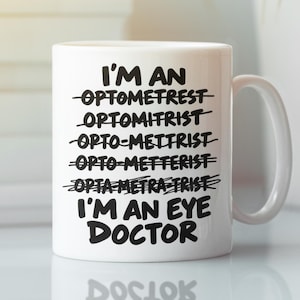 Op de afbeelding: Een witte keramische mok met zwarte tekst die luidt I'M AN OPTOMETREST, OPTOMITRIST, OPTO-METTRIST, OPTO-METTERIST, OPTA METRA TRIST, allemaal doorgestreept met horizontale lijnen, gevolgd door de duidelijke tekst I'M AN EYE DOCTOR.
