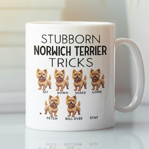 以下が含まれることがあります： 「STUBBORN NORWICH TERRIER TRICKS」の文字と、ノリッチ・テリア犬がトリックをしているイラストが描かれた白いセラミックマグカップ。トリックには、座る、伏せる、振る、来る、持ってくる、転がる、待てが含まれます。マグカップには白いハンドルが付いています。