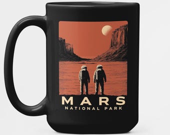 Op de afbeelding: Zwarte keramische mok met een Mars National Park ontwerp. De afbeelding toont twee astronauten op een rode planeet, met een volle maan en een canyonlandschap. De mok heeft een groot handvat.