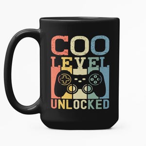 Puede incluir: Taza negra con la frase "COO LEVEL UNLOCKED" en una fuente retro, junto con un gráfico de un mando de videojuegos. El texto y el mando son en tonos rojos, amarillos y azules.