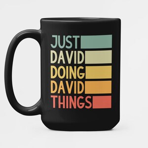 Puede incluir: Taza de cerámica negra con el texto "JUST DAVID DOING DAVID THINGS" en un diseño retro de bloques de color. La taza tiene un asa grande y un diseño sencillo y moderno.