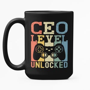 Puede incluir: Taza de cerámica negra con las palabras "CEO LEVEL UNLOCKED" en una fuente retro, junto con un gráfico de mando colorido. La taza tiene un asa grande y un fondo blanco.