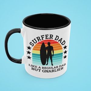 May include: White ceramic coffee mug with a black rim and handle. The mug features a retro-style graphic of a surfer with a surfboard and the text "Surfer Dad Like a Regular Dad But Gnarlier".