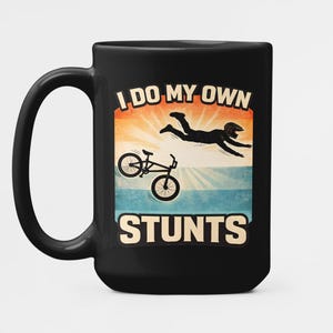 以下が含まれることがあります： 「I DO MY OWN STUNTS」の文字とBMXバイクとスタントをしている人のグラフィックが描かれた黒いセラミックマグカップ。デザインは、オレンジ、黄色、青の色合いの夕焼けのような背景が特徴です。