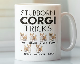 May include: White ceramic coffee mug featuring a black graphic of a corgi dog and the text "Stubborn Corgi Tricks". The graphic depicts a corgi dog in various poses with the text "Sit", "Down", "Shake", "Come", "Fetch", "Roll Over", and "Stay" beneath each pose.