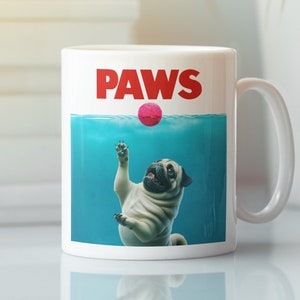 Może przedstawiać: Biały kubek ceramiczny z niebiesko-białym obrazkiem mopsa pod wodą, który sięga po różową piłkę. Tekst "PAWS" jest nadrukowany na czerwono nad obrazkiem.