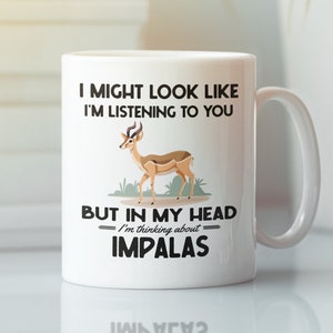 Op de afbeelding: Een witte keramische koffiemok met een zwart-bruine illustratie van een impala. De tekst op de mok luidt "I might look like I'm listening to you, but in my head I'm thinking about impalas."
