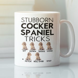 Könnte beinhalten: Weiße Keramik-Kaffeetasse mit einer schwarzen Grafik eines Cockerspaniels und dem Text "Stubborn Cocker Spaniel Tricks". Die Grafik zeigt einen Cockerspaniel in verschiedenen Posen mit dem Text "Sit", "Down", "Shake", "Come", "Fetch", "Roll Over" und "Stay" darunter.
