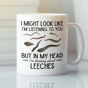 Puede incluir: Taza de cerámica blanca con un gráfico negro que dice "Puedo parecer que te estoy escuchando, pero en mi cabeza estoy pensando en sanguijuelas". El gráfico incluye una serie de sanguijuelas de dibujos animados.