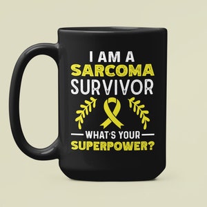 以下が含まれることがあります： 白いグラフィックで「私は肉腫の生存者です」と書かれた黒いコーヒーカップ。黄色のリボンと「あなたのスーパーパワーは何ですか？」というテキストがあります。