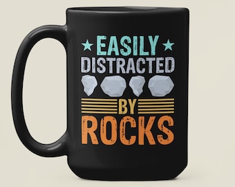 May include: Black coffee mug with white text that reads "Easily Distracted By Rocks" with a graphic of five rocks in a row.