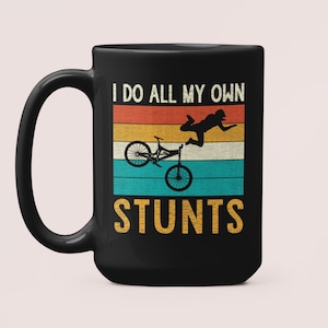 May include: Black coffee mug with a white graphic of a person doing a bike stunt. The graphic includes a vintage-style rainbow stripe background and the text "I do all my own stunts".