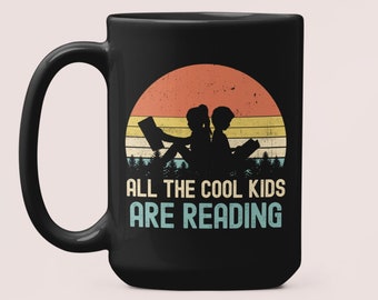 May include: Black coffee mug with a retro sunset design and the text "All the cool kids are reading". The design features two children sitting and reading books.