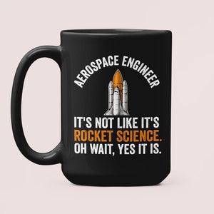 Op de afbeelding: Zwarte keramische koffiemok met een witte afbeelding van een spaceshuttle en de tekst "AEROSPACE ENGINEER IT'S NOT LIKE IT'S ROCKET SCIENCE. OH WAIT, YES IT IS."