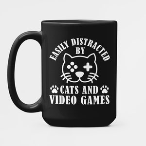 May include: Black coffee mug with white text that reads "Easily Distracted By Cats And Video Games". A cartoon cat with a video game controller is in the center of the text.