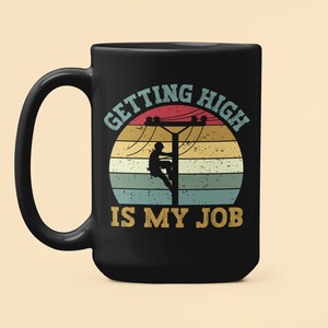 Puede incluir: Taza de café negra con un gráfico de estilo vintage de una persona escalando un poste de energía. El texto dice "Getting High Is My Job".