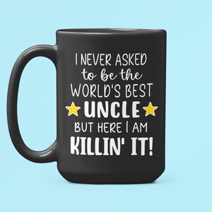 May include: Black ceramic mug with the text "I NEVER ASKED to be the WORLD'S BEST UNCLE BUT HERE I AM KILLIN' IT!" in white lettering. The word "UNCLE" is between two yellow stars. The mug has a large handle.