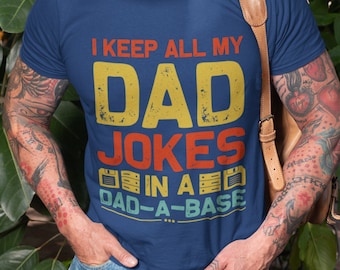 May include: A man wearing a blue t-shirt with the text "I keep all my dad jokes in a dad-a-base" printed on it. The text is in a retro style font with a yellow, red, and blue color scheme. The man is smiling and has a white beard.