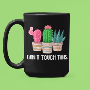 以下が含まれることがあります： 白い取っ手付きの黒いセラミック製のコーヒーカップ。カップには、4つの鉢植えのサボテンと「Can't Touch This」という文字が書かれたグラフィックが描かれています。