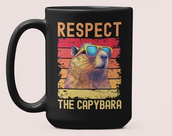 Op de afbeelding: Zwarte koffiemok met een afbeelding van een capybara met een zonnebril. De afbeelding is in retro-stijl met een rood, oranje en geel kleurenschema. De tekst "RESPECT THE CAPYBARA" staat onder de afbeelding.