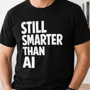 May include: A black short-sleeved t-shirt featuring the white text "STILL SMARTER THAN AI" printed in a bold, sans-serif font across the chest. The garment has a classic crew neckline and a straight hem, constructed from a solid black fabric.