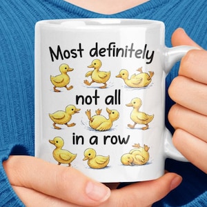 May include: White ceramic mug featuring yellow cartoon ducklings and the text "Most definitely not all in a row." The mug has a white handle and is held against a blue background.