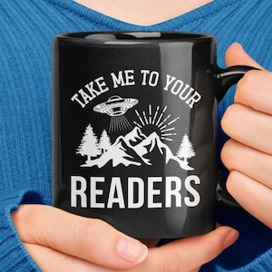 Pode incluir: Caneca de café preta com um design branco de um OVNI, montanhas e árvores. O texto na caneca diz "Take me to your readers".